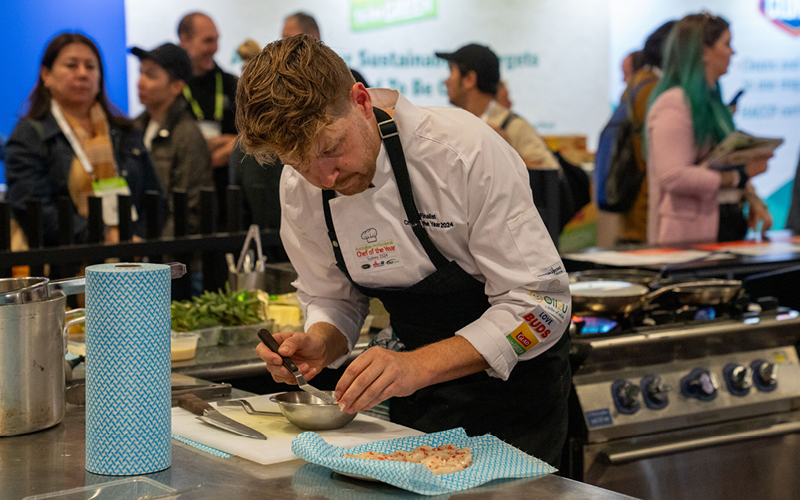 Chef in a white coat and black apron preparing food in a professional kitchen competing at chef of the year