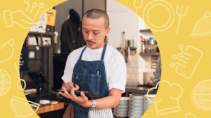 A chef wearing a denim apron uses a digital tablet inside a busy commercial kitchen, surrounded by plates and equipment, with a yellow hospitality-themed graphic overlay framing the scene.