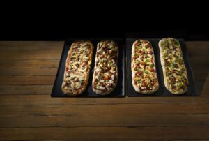 Four rectangular pizzas with various toppings on black trays placed on a wooden table.