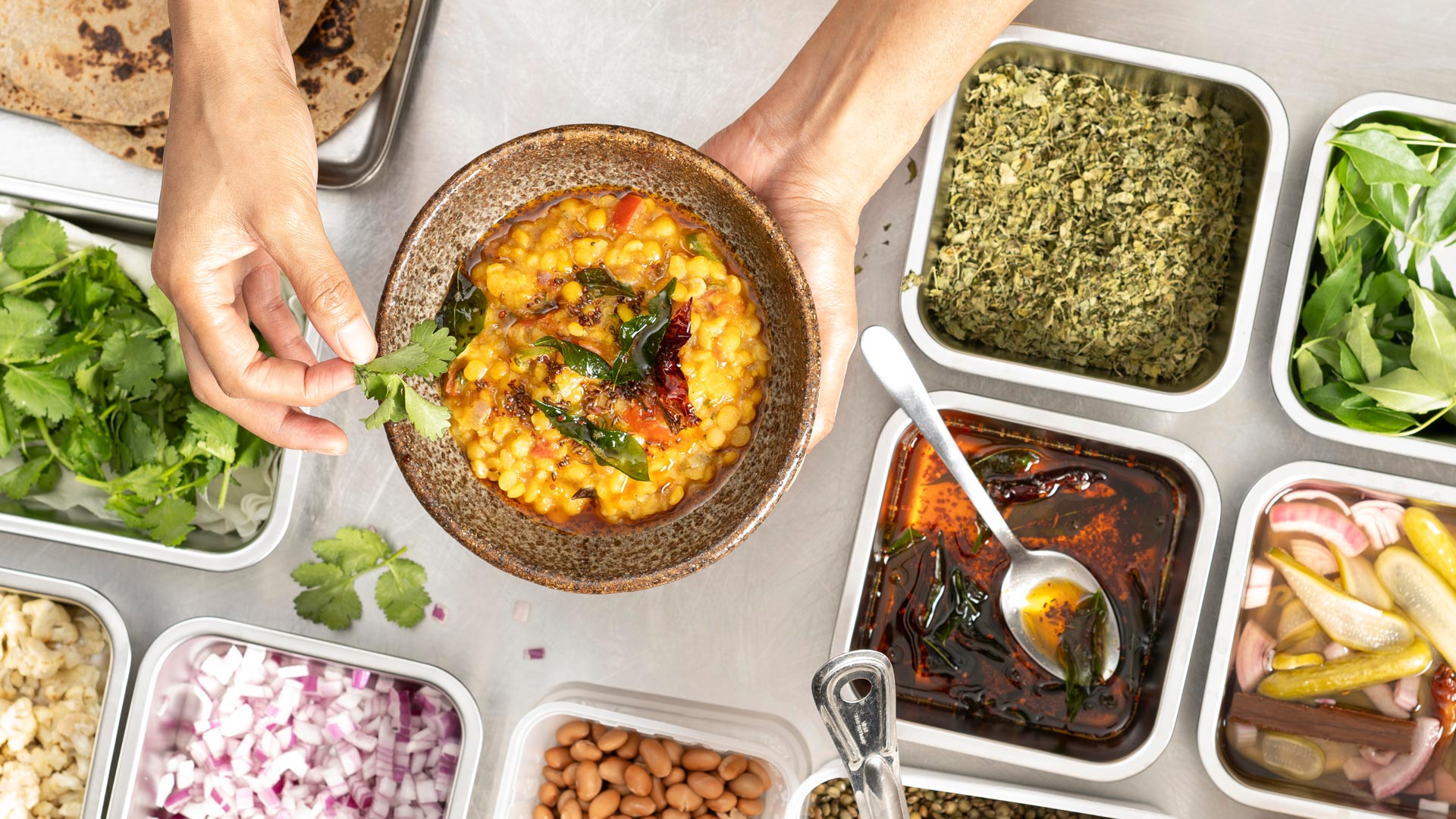 A top-down view of a person holding a bowl of food, known as Indian Dal Tadka. The dish is garnished with fresh herbs and spices, surrounded by various ingredient containers on a rustic wooden surface.