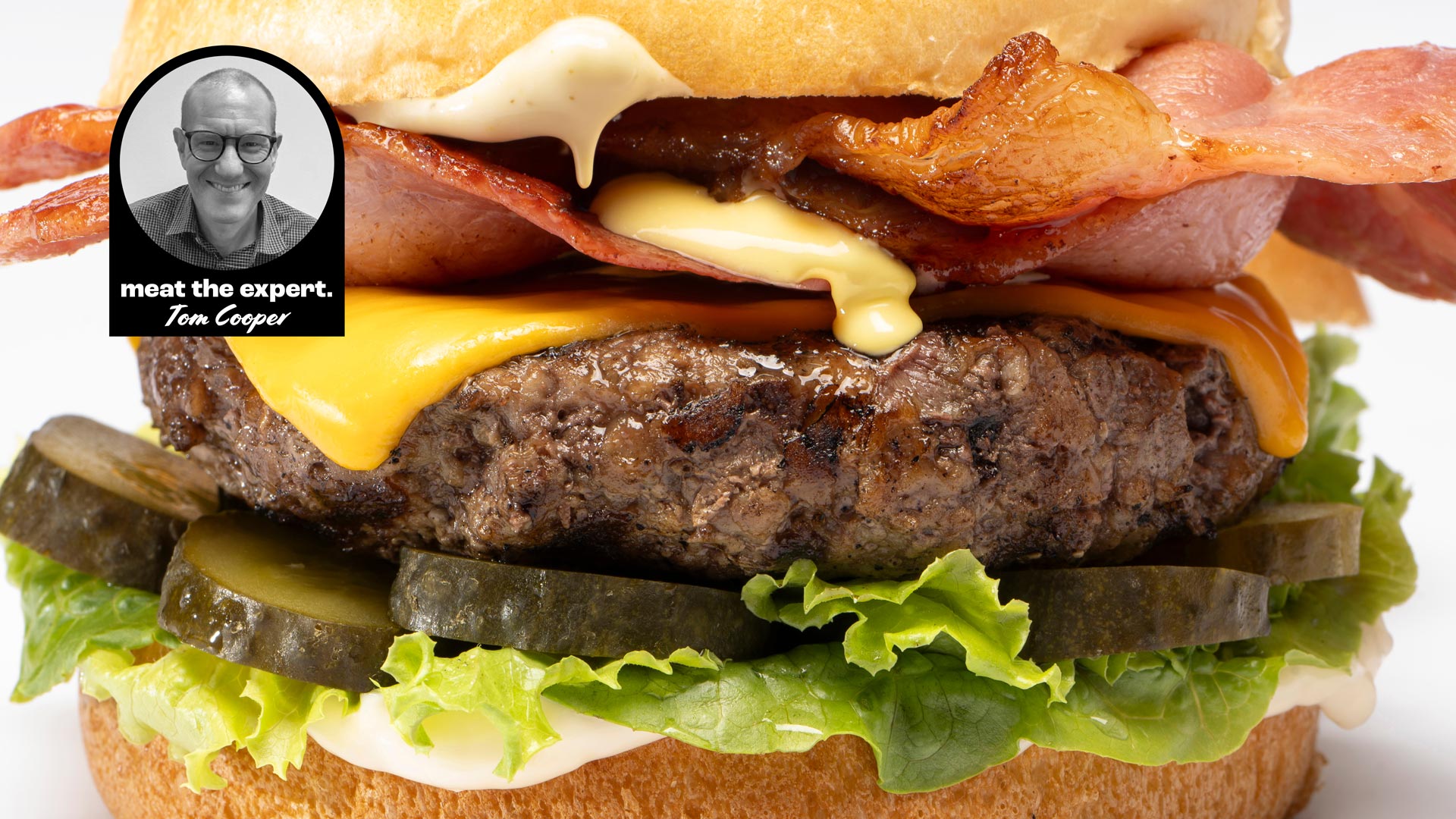 Close-up of a hamburger with lettuce, pickles, a beef patty, melted cheese, bacon, and sauce, with a label featuring Tom Cooper and the text "meat the expert. Tom Cooper."