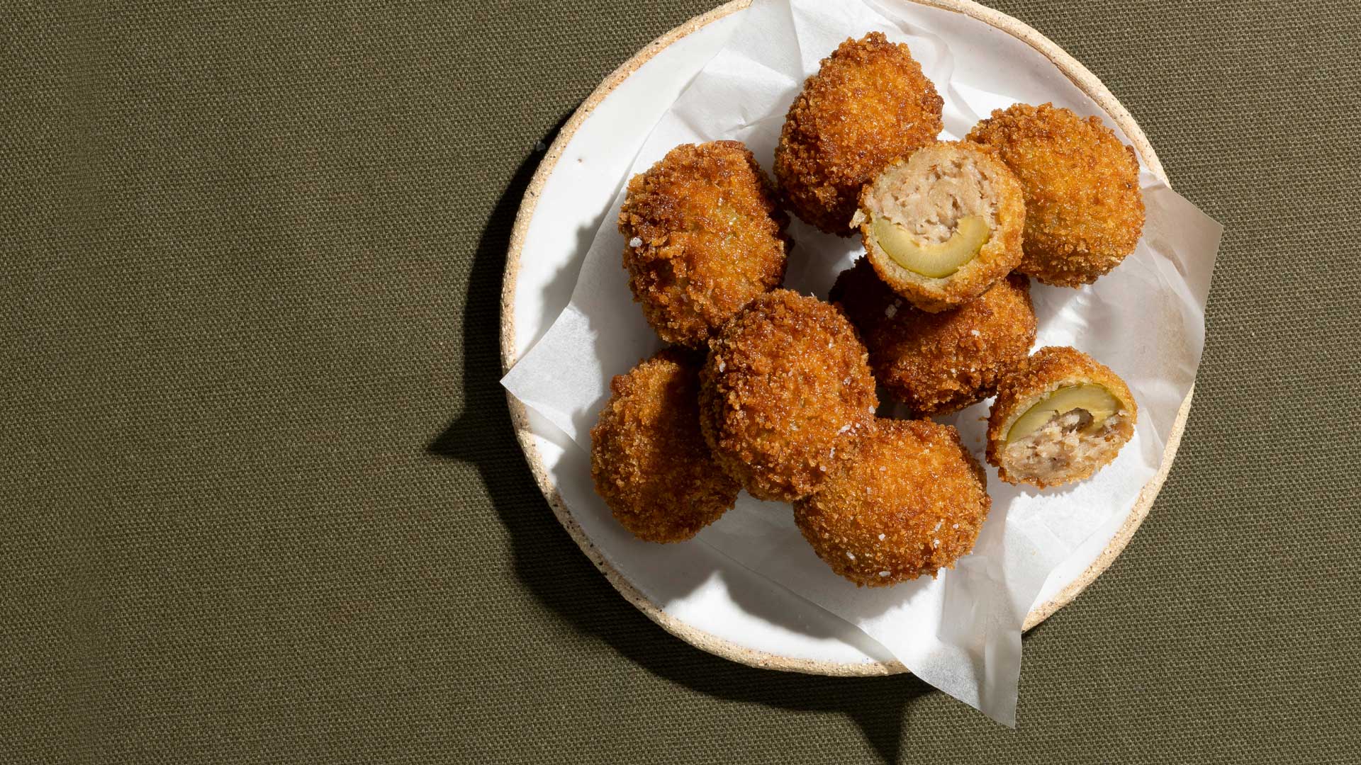 A plate of fried stuffed olives, known as Olive all’Ascolana, on a green textured surface. The olives are breaded and fried, with one olive cut in half to show the stuffing inside.