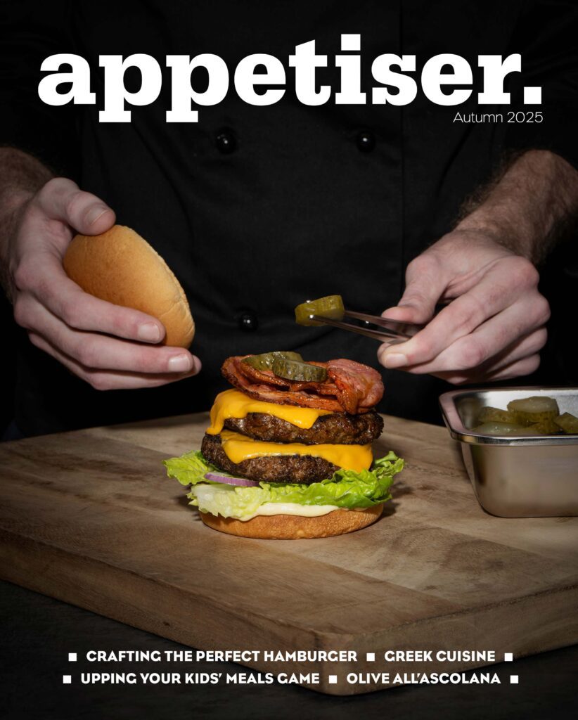 A person assembling a hamburger with multiple layers of ingredients on a wooden board, with the title "appetiser." and the subtitle "Autumn 2025" at the top. The bottom of the image lists article titles: "Crafting the Perfect Hamburger," "Greek Cuisine," "Upping Your Kids' Meals Game," and "Olive All'Ascolana."