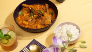 A bowl of Thai red curry with roast duck garnished with herbs and sliced chilli, served alongside steamed jasmine rice and a refreshing cucumber cocktail on a yellow tablecloth.