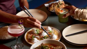 Diners share a Southeast Asian feast, with cocktails, rice and vibrant plates of food including coconut-topped fish and herb-laced dishes on a restaurant table.
