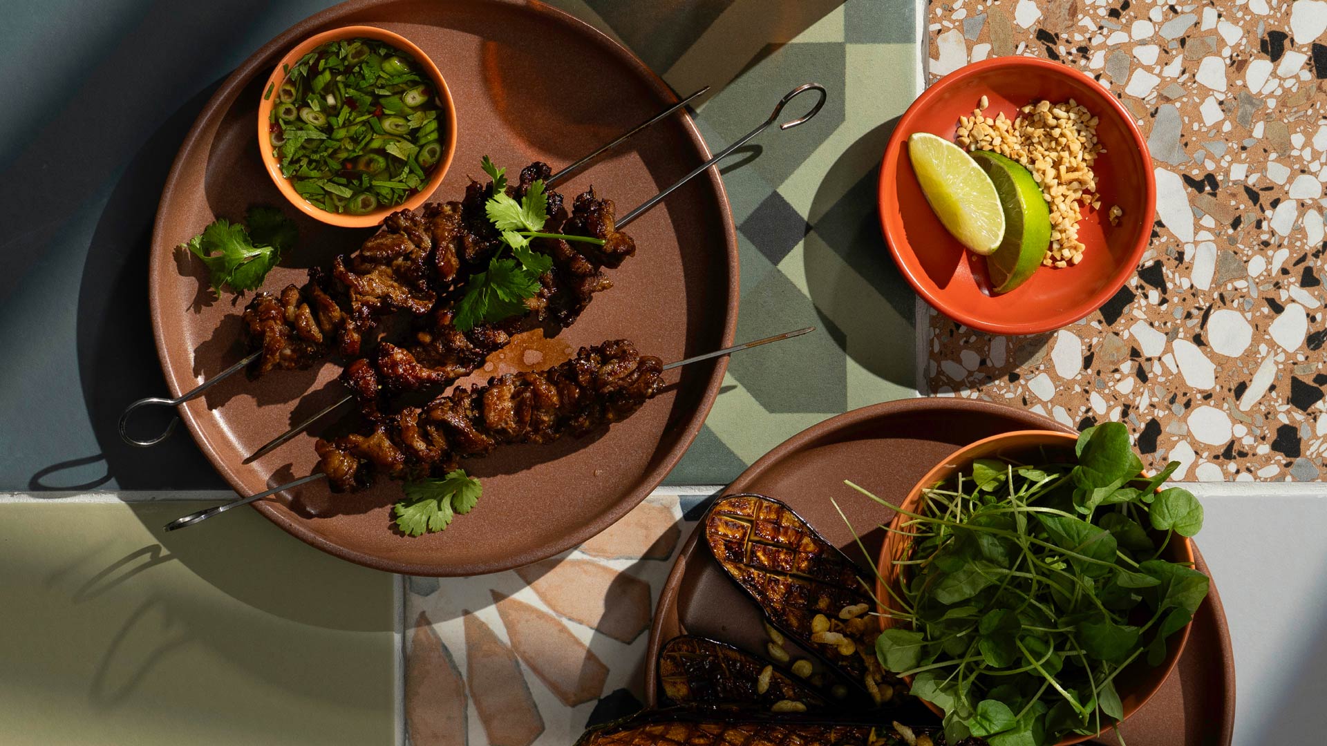 Moo ping — Thai grilled pork skewers served with dipping sauce, fresh herbs, charred eggplant, lime wedges, and crushed peanuts on terracotta plates.