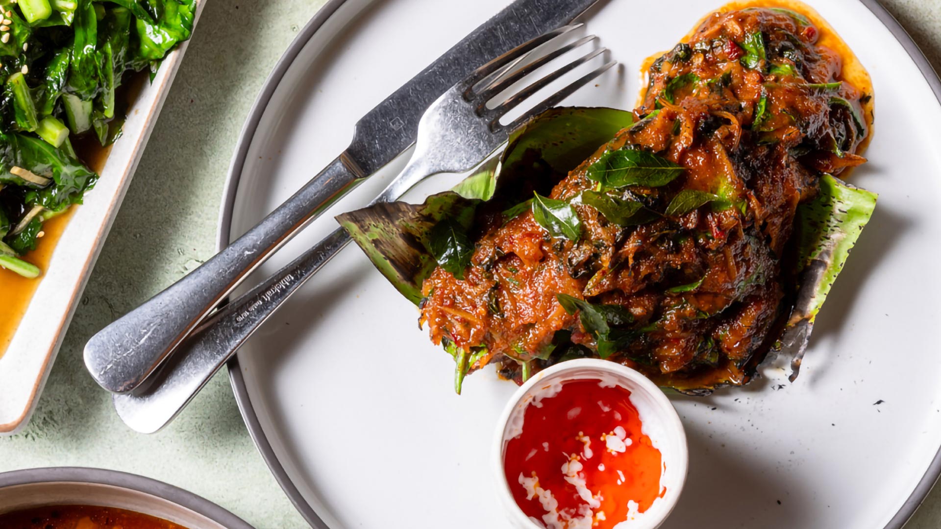 Grilled salmon wrapped in banana leaf, topped with a rich red chilli paste and fresh herbs, served with a small cup of bright red chilli dipping sauce.