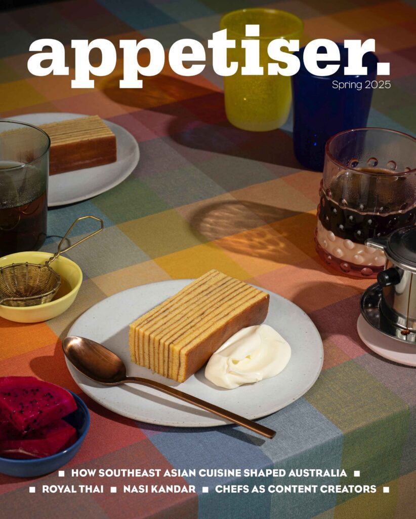 Cover of appetiser magazine, Spring 2025 edition. A slice of Indonesian layer cake (kue lapis legit) served with cream on a white plate with a bronze spoon, dragon fruit, tea, and colourful glassware on a chequered tablecloth.