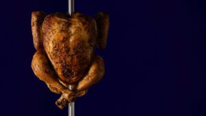 Whole rotisserie chicken cooked golden brown on a spit against a dark blue background.