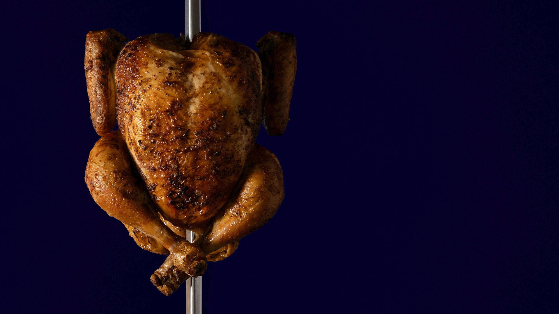 Whole rotisserie chicken cooked golden brown on a spit against a dark blue background.
