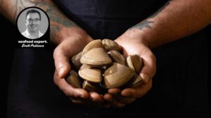 Tattooed hands holding a large handful of fresh Cloudy Bay clams, with a photo inset of seafood expert Brett Patience.