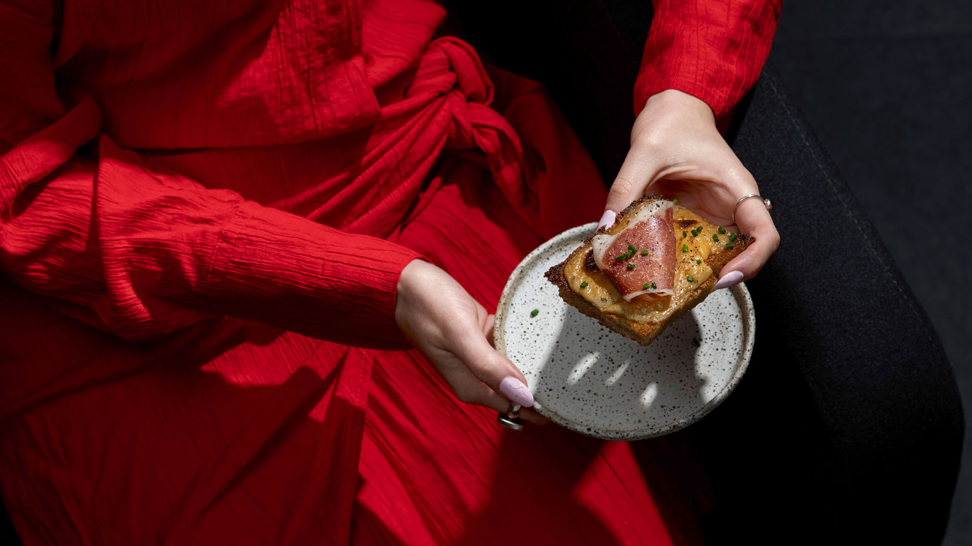 A woman in a red dress holding a ceramic plate with a slice of Welsh rarebit topped with cured meat and chives.
