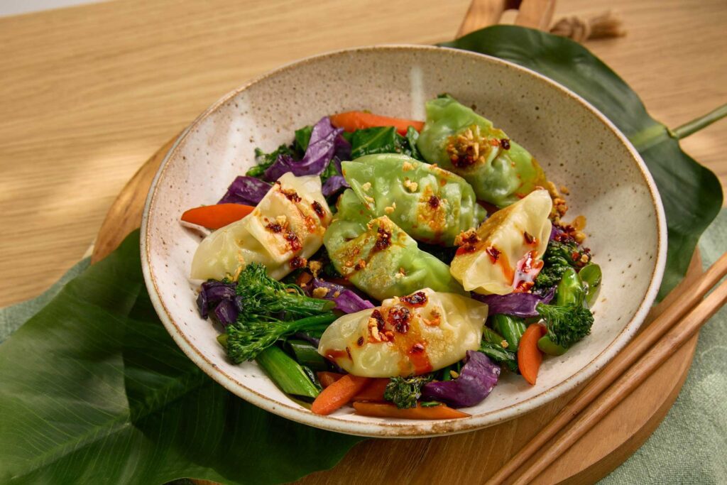 Appetising dish featuring dumplings served over a colourful stir-fry. The dumplings are topped with a drizzle of chilli oil and sprinkled with crispy garlic. Beneath them is a mix of fresh vegetables, including broccolini, purple cabbage, carrot, and green beans, all lightly stir-fried to retain their bright colours and textures.