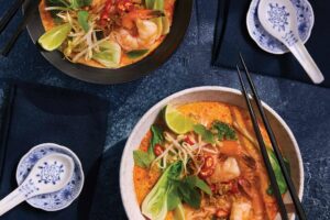 Two bowls of laksa curry, topped with fresh herbs, lime, bok choy, and chilies, are beautifully presented with chopsticks and porcelain spoons on a dark textured surface.