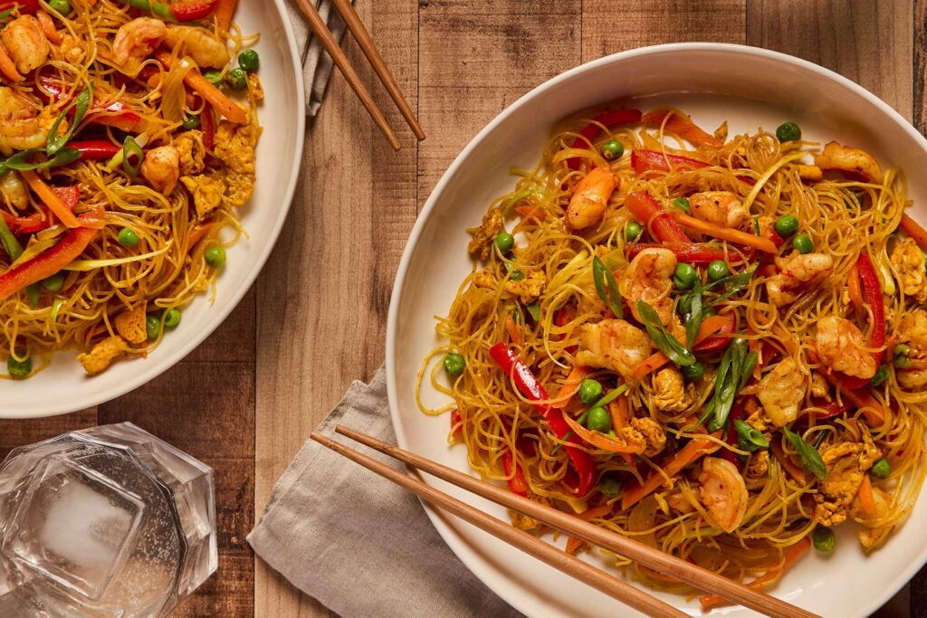 Two bowls of Singapore noodles on a table with chopsticks and a napkin. There is also two glasses of soft drink with ice in it.