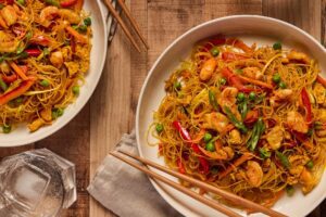 Two bowls of Singapore noodles on a table with chopsticks and a napkin. There is also two glasses of soft drink with ice in it.