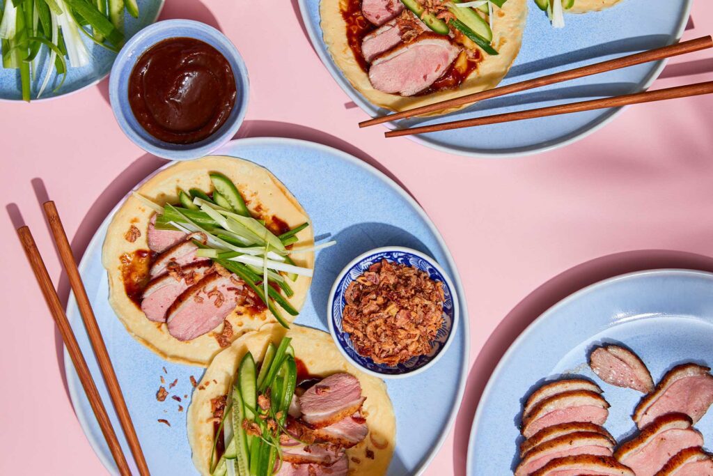 A close-up image of a delicious peking duck pancakes being assembled. The soft, warm pancake is topped with tender slices of roasted duck, a drizzle of hoisin sauce, and garnished with fresh cucumber and scallions. The pancake is ready to be eaten, with a few more pancakes and ingredients visible in the background.