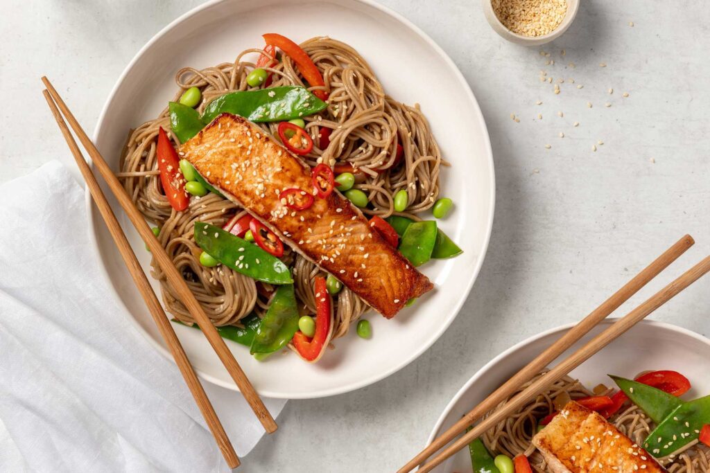 Two bowls of soba noodle salad with salmon. Each bowl contains soba noodles mixed with vegetables like snap peas, red capsicum, and edamame. On top of the noodles sits a piece of glazed salmon, garnished with sesame seeds and sliced red chili.